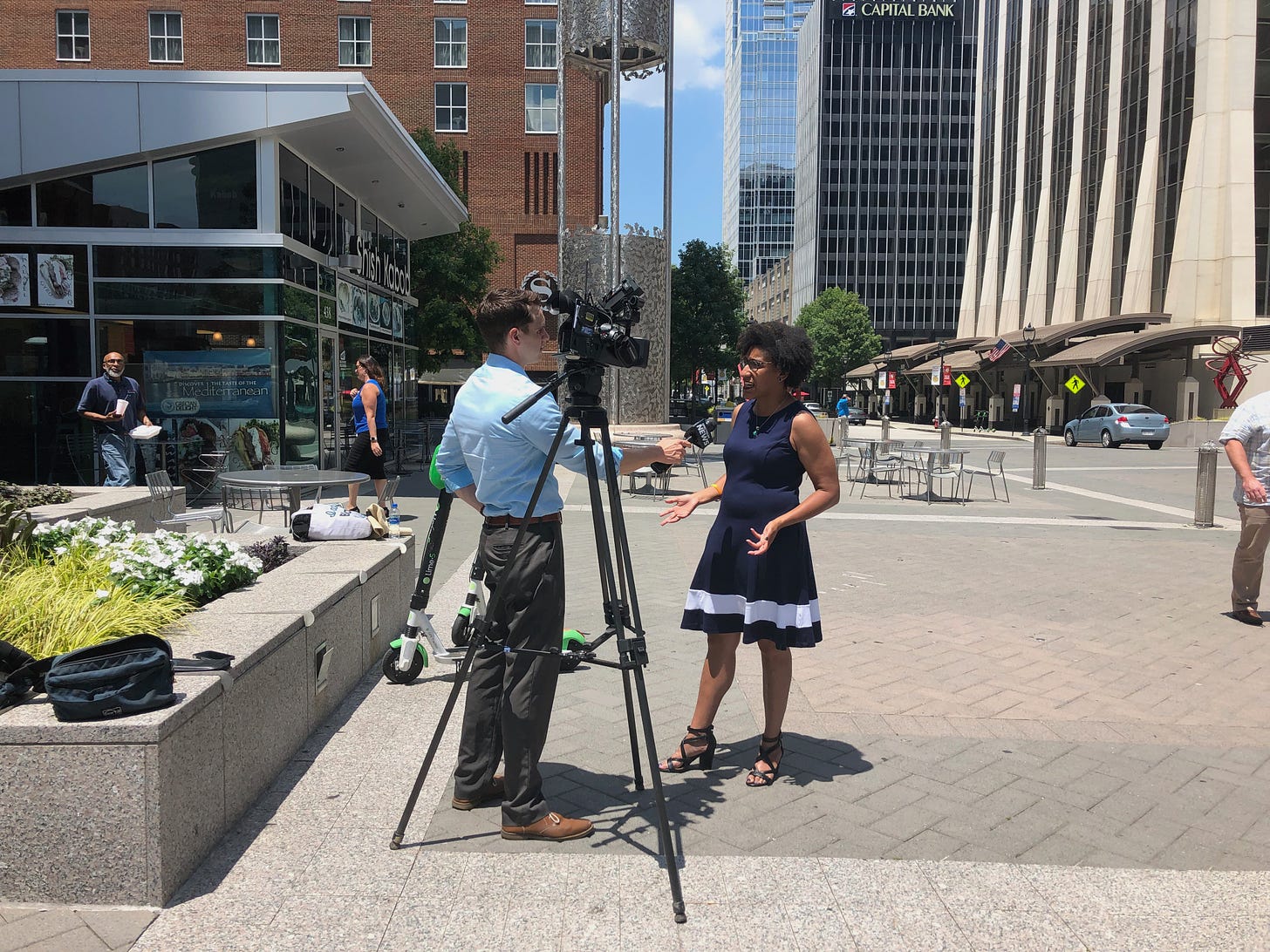 Kristen is wearing a blue dress with a white stripe and she's standing next to a reporter in a blue shirt and green kakhis and their camera on the Fayetteville Street Mall in Downtown Raleigh, NC in June of 2019