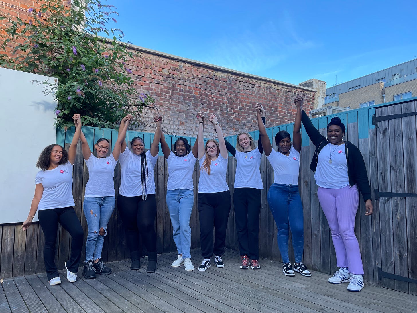 The London team standing outside, arms in the air and holding hands, wearing matching white t-shirts with the girls' project logo on them