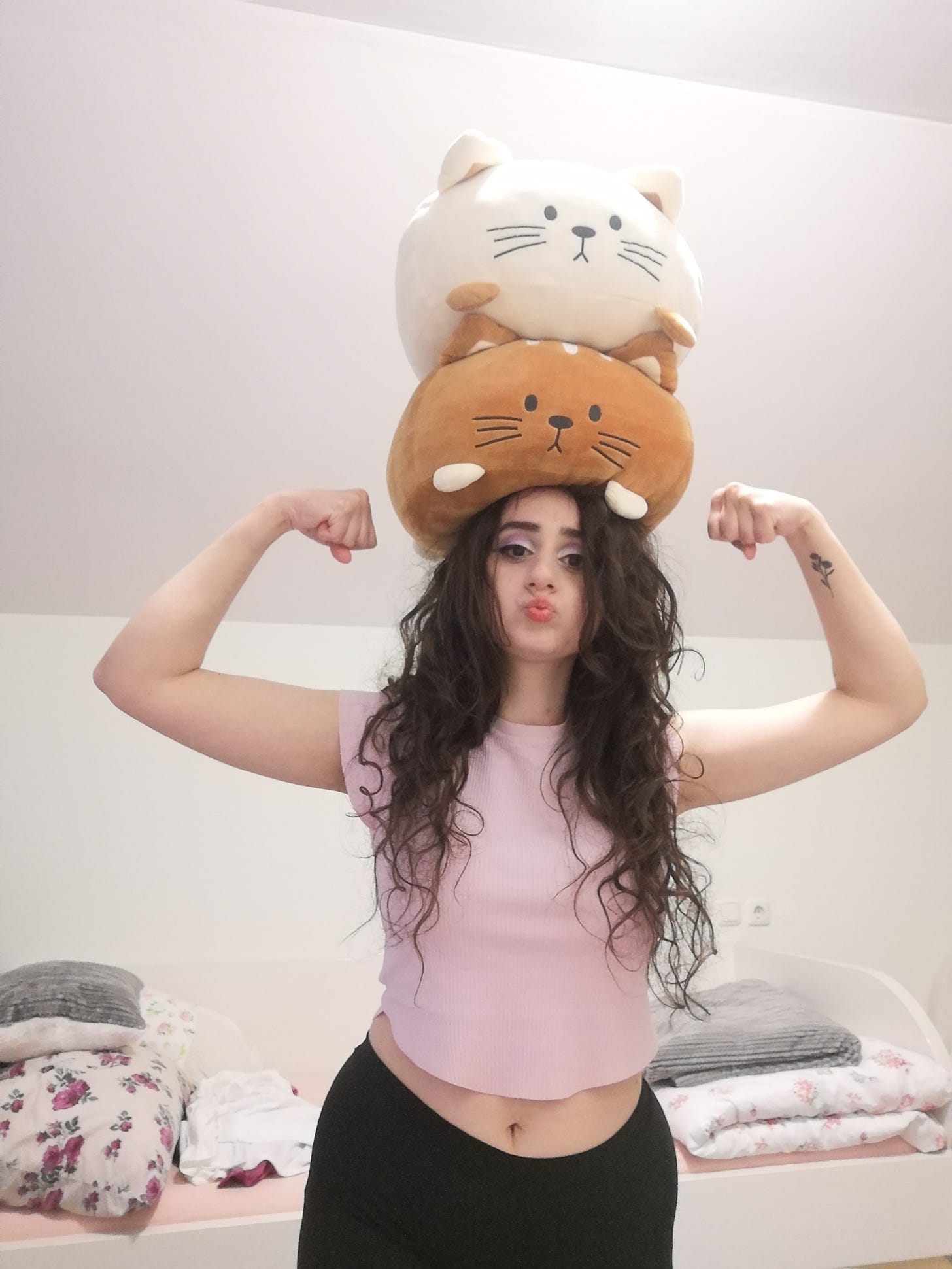 Matea, a girl with dark-brown, bust-length hair, puckering her lips and flexing her arms while balancing two cat plushies on her head