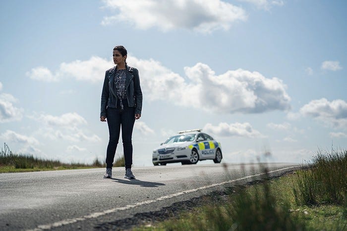 This image shows Yasmin "Yaz" Khan (played by Mandip Gill) standing on a road in the English countryside, on a bright and clear day, in "Can You Hear Me?" A police cruiser sits on the road several feet behind Yaz. This image shows Yasmin "Yaz" Khan (played by Mandip Gill) standing on a road in the English countryside, on a bright and clear day, in "Can You Hear Me?" A police cruiser sits on the road several feet behind Yaz.