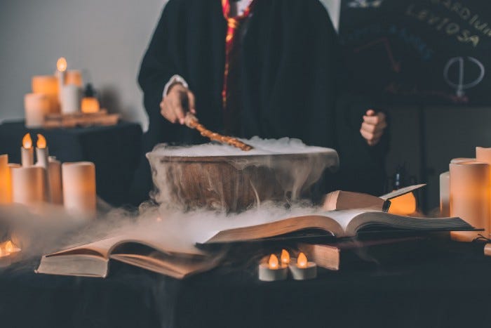 A person dressed in wizard robes holding a wand over a foggy cauldron. There are books, candles, and more laid out, making it seem like a magical/fantasy setting.