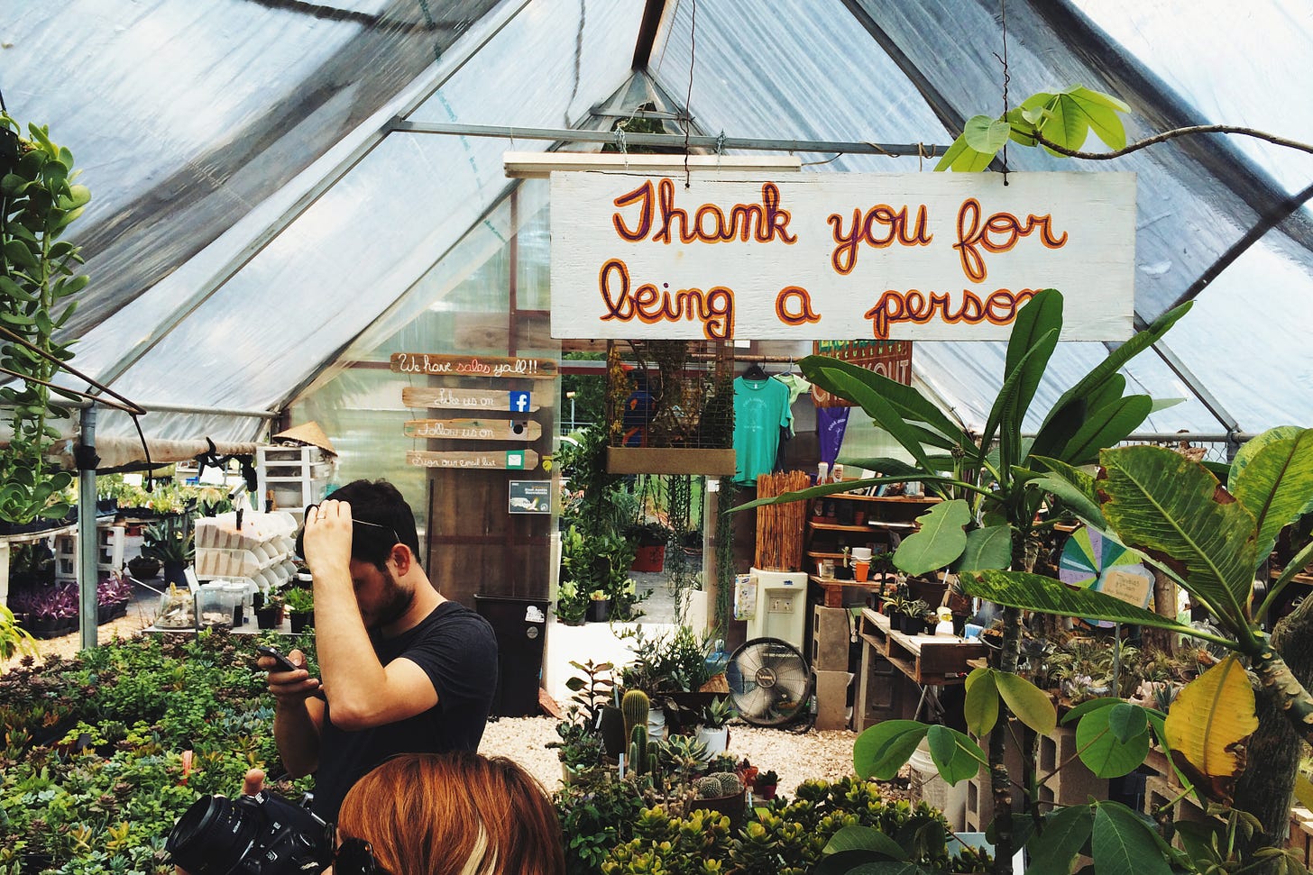 Two people shopping in a plant store, a sign hangs over them that reads Thank You For Being a Person.