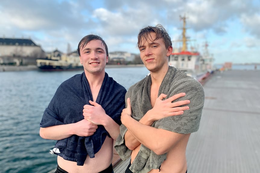 Two men smile while holding towels over their shoulders after a swim.