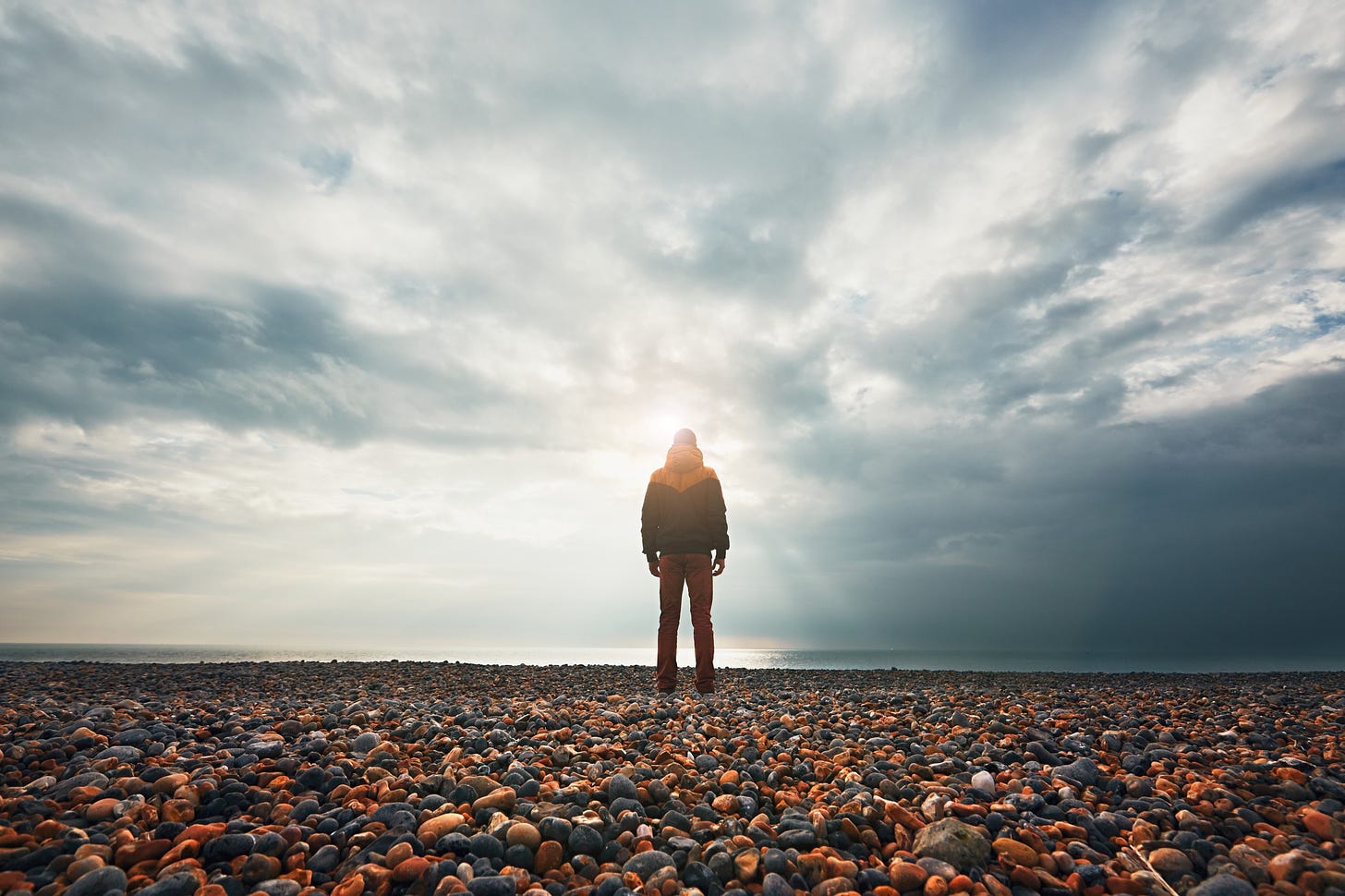 A person stands alone on a rocky beach and stares out toward the sea and endless sky.