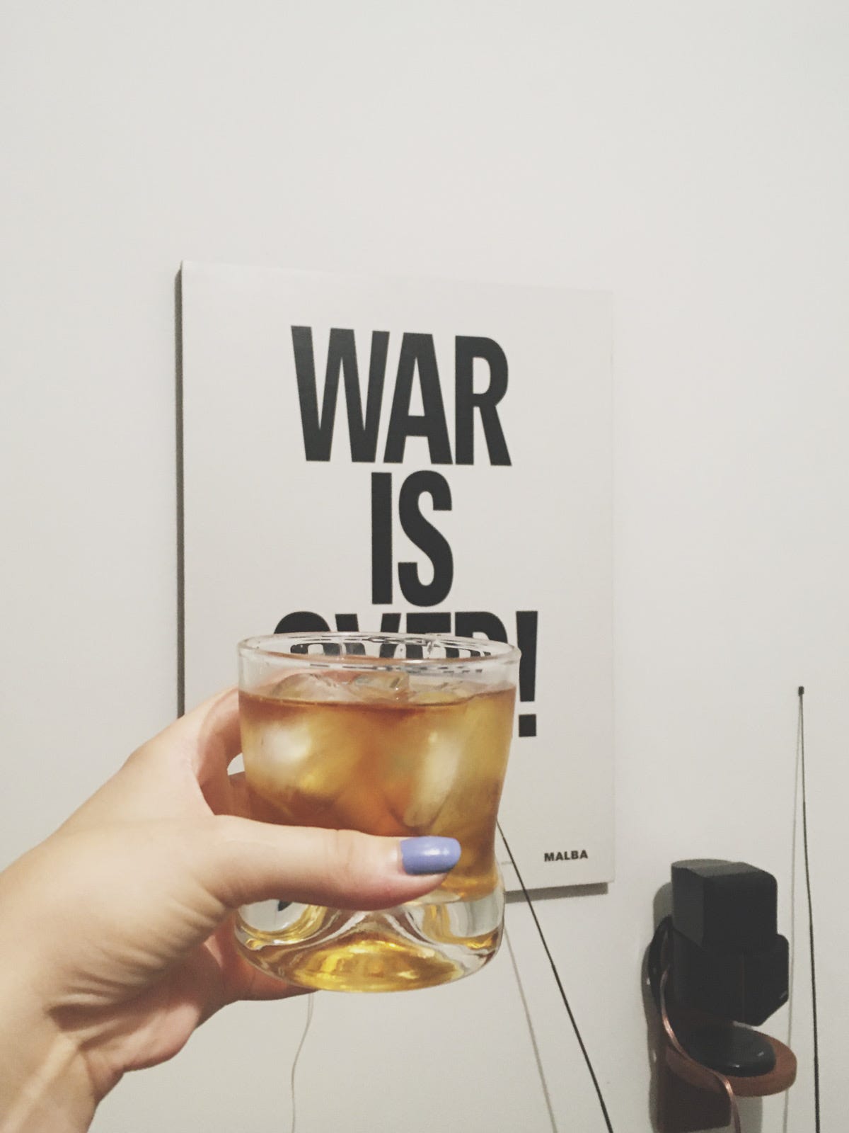 Foto em que estou com o braço estendido segurando um copo de uísque e ao fundo tem um poster na parede escrito WAR IS OVER