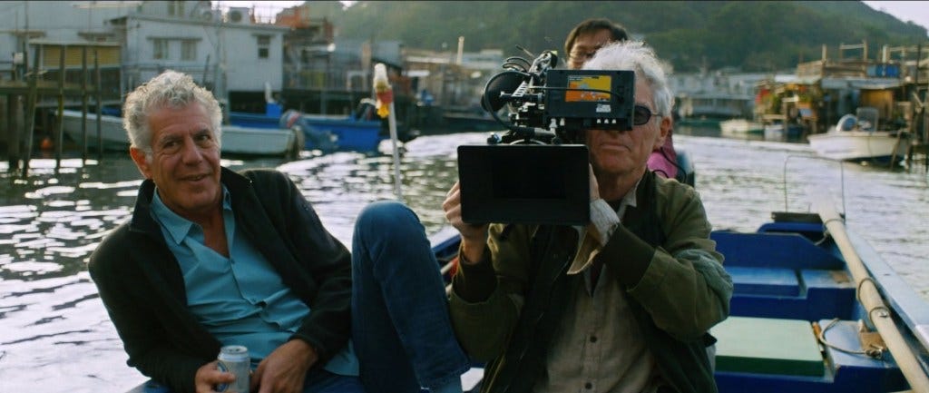 Anthony Bourdain and Christopher Doyle sit on a boat in Hong Kong. Doyle is holding a large camera.