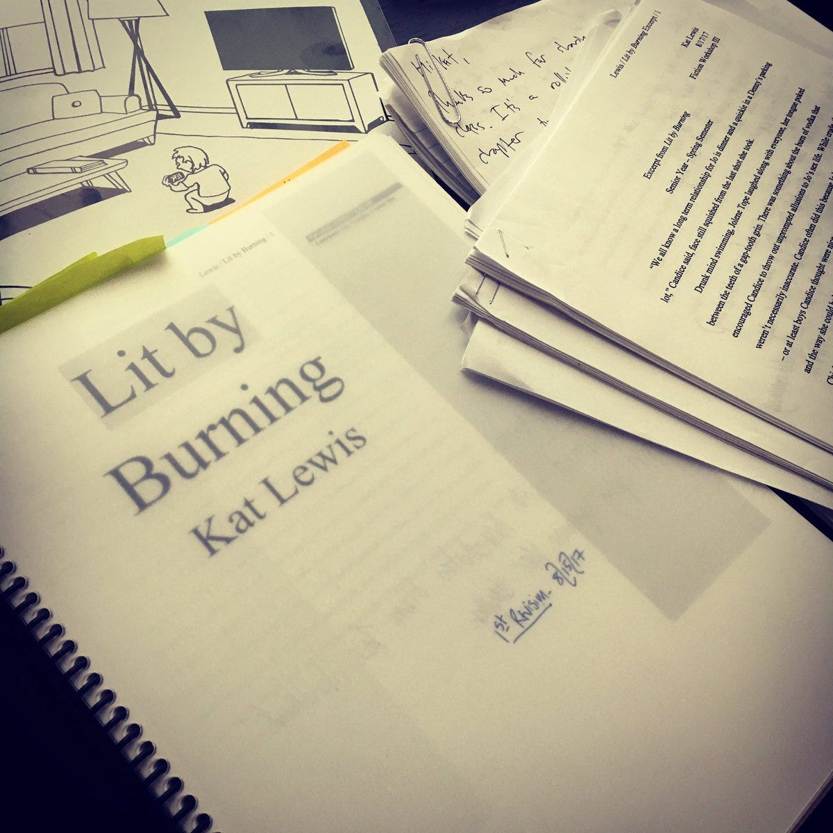 A photo of Kat’s messy writing desk. There is a bound print out of her novel, Lit by Burning, which is dated August 15, 2017.