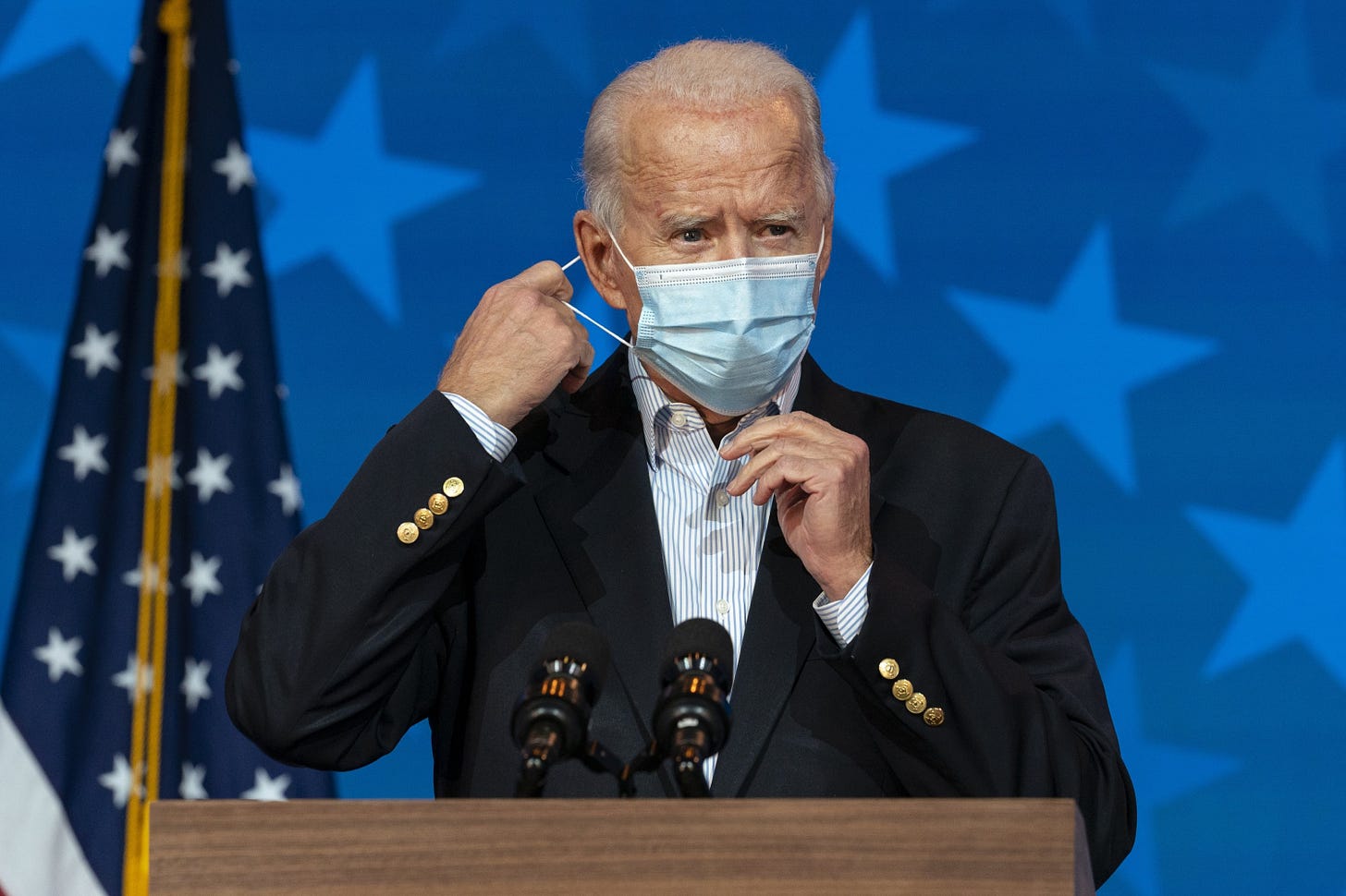 President Joe Biden stands in front of a podium. He is removing a surgical mask from his face. The background is blue with white stars, and an American flag hangs behind him. The look on Biden's face is stern.