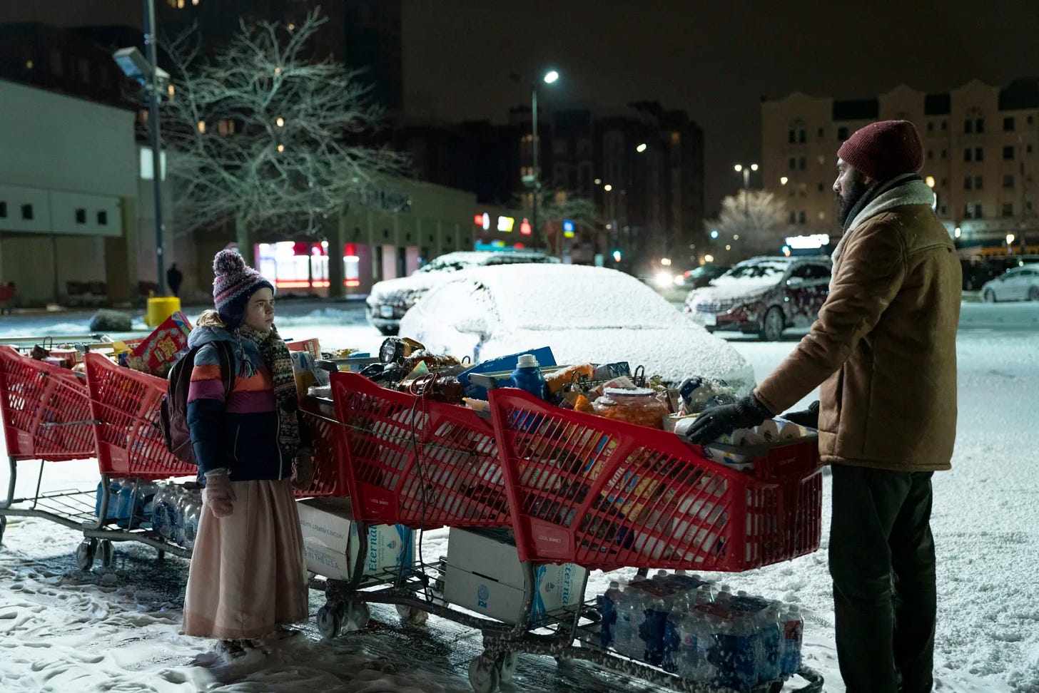 Matilda Lawler and Himesh Patel standing out in a snowy supermarket carpark with a line of trolleys stacked with groceries in Station Eleven.