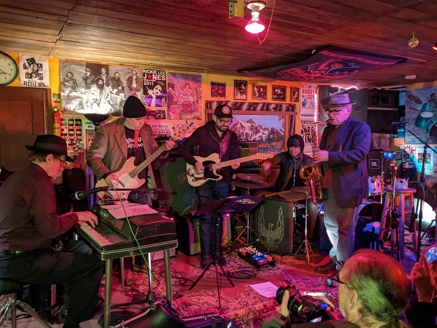 A five piece band of keyboard, bass, guitar, drums, and sax, playing in a small room filled with music memorabilia