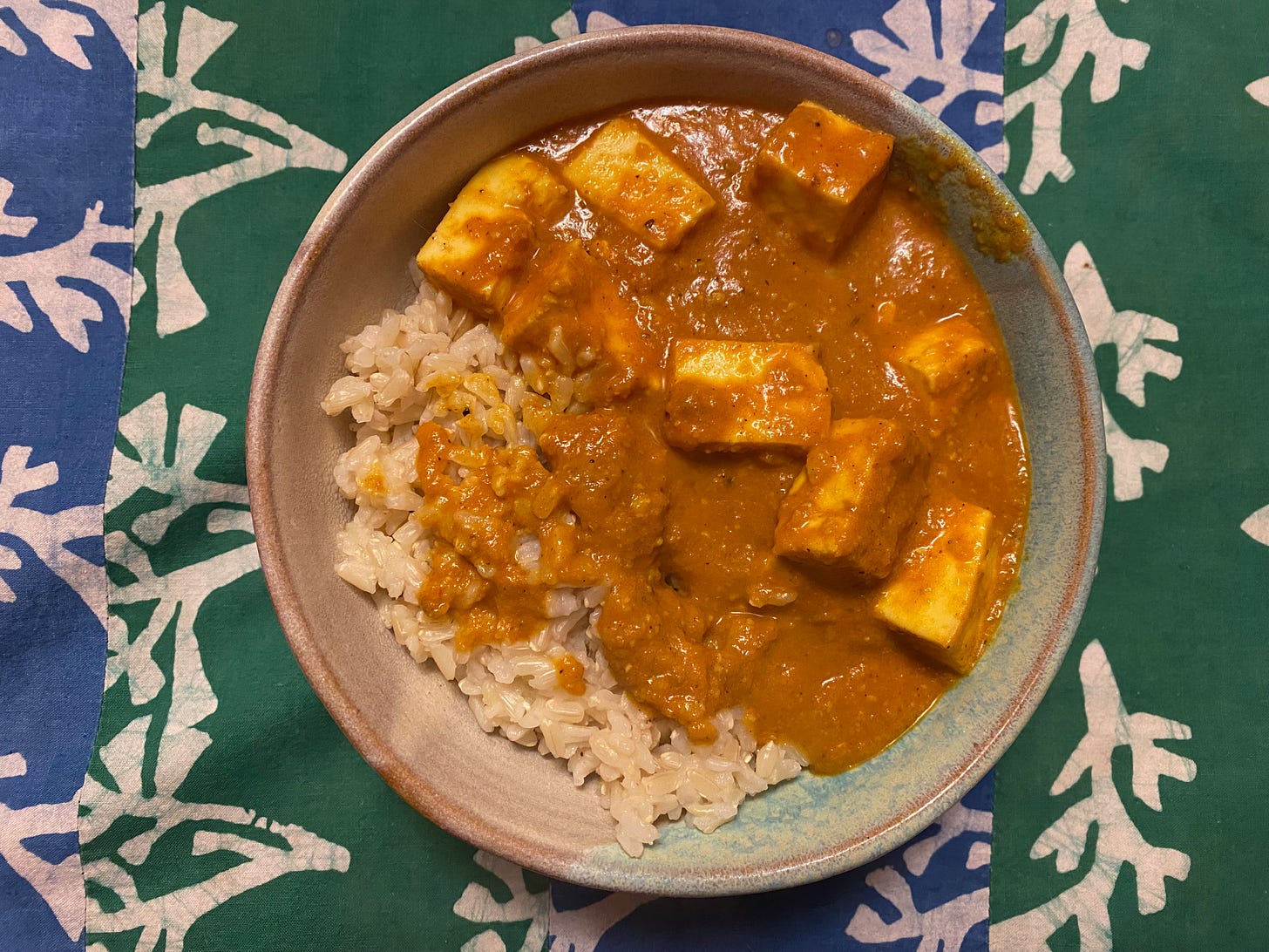 A ceramic bowl of rice and curry sits on a blue and green striped placemat. The deep orange sauce is thick and creamy, with large chunks of paneer.