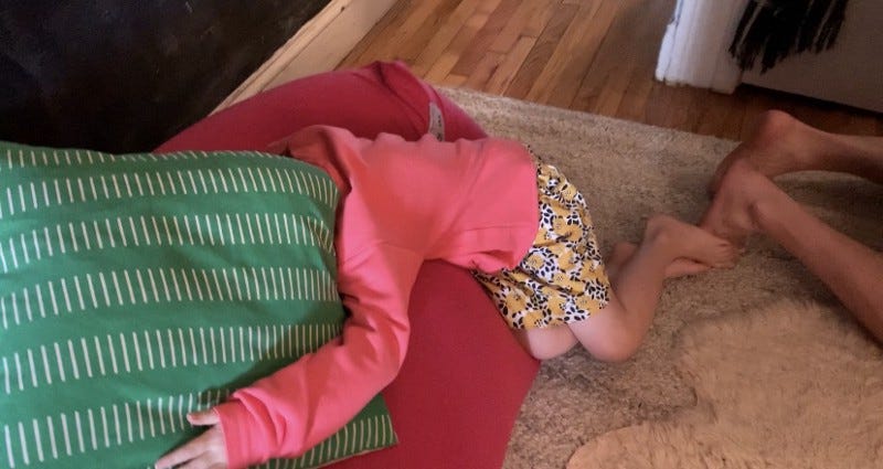 A kid buries her head under a pillow, embarrassed by her parents. She wears a coral colored sweatshirt and a floral skirt.