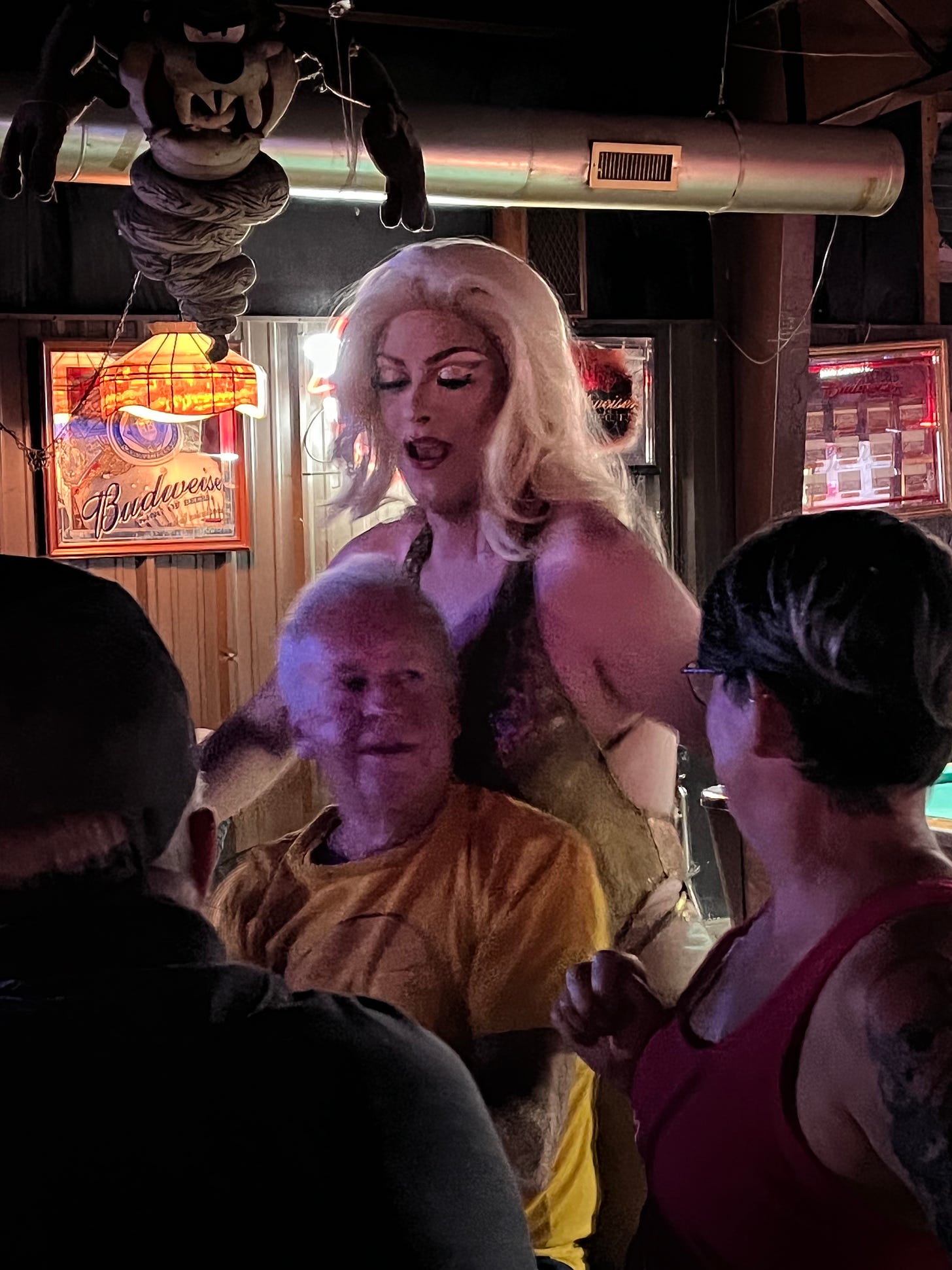 A drag queen with blonde hair and a sparkly top stands behind an older male bar patron and flirts with him.
