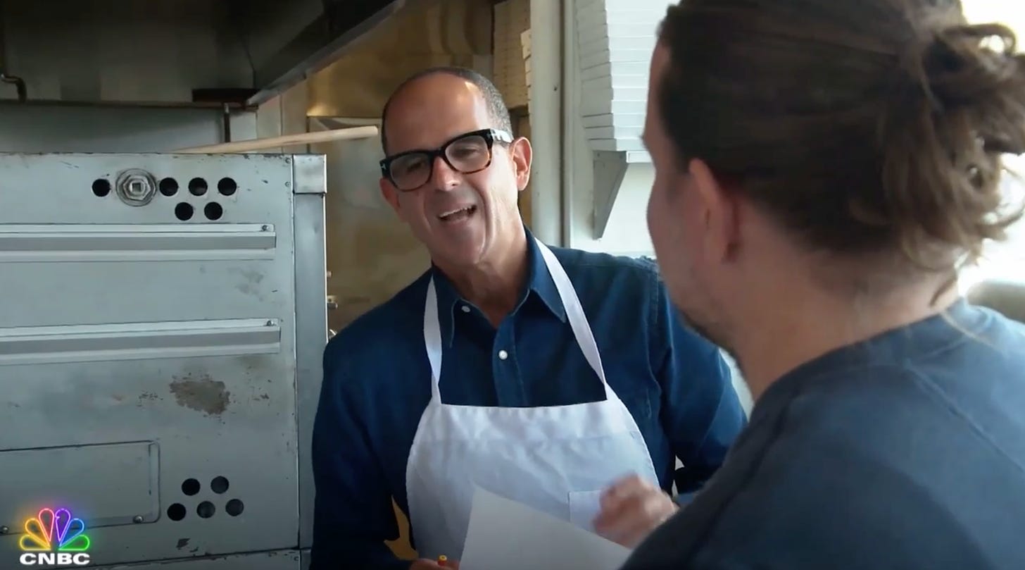 screen shot of tv show the profit with man in apron and piece of paper looking irritated at man with man bun standing in foreground