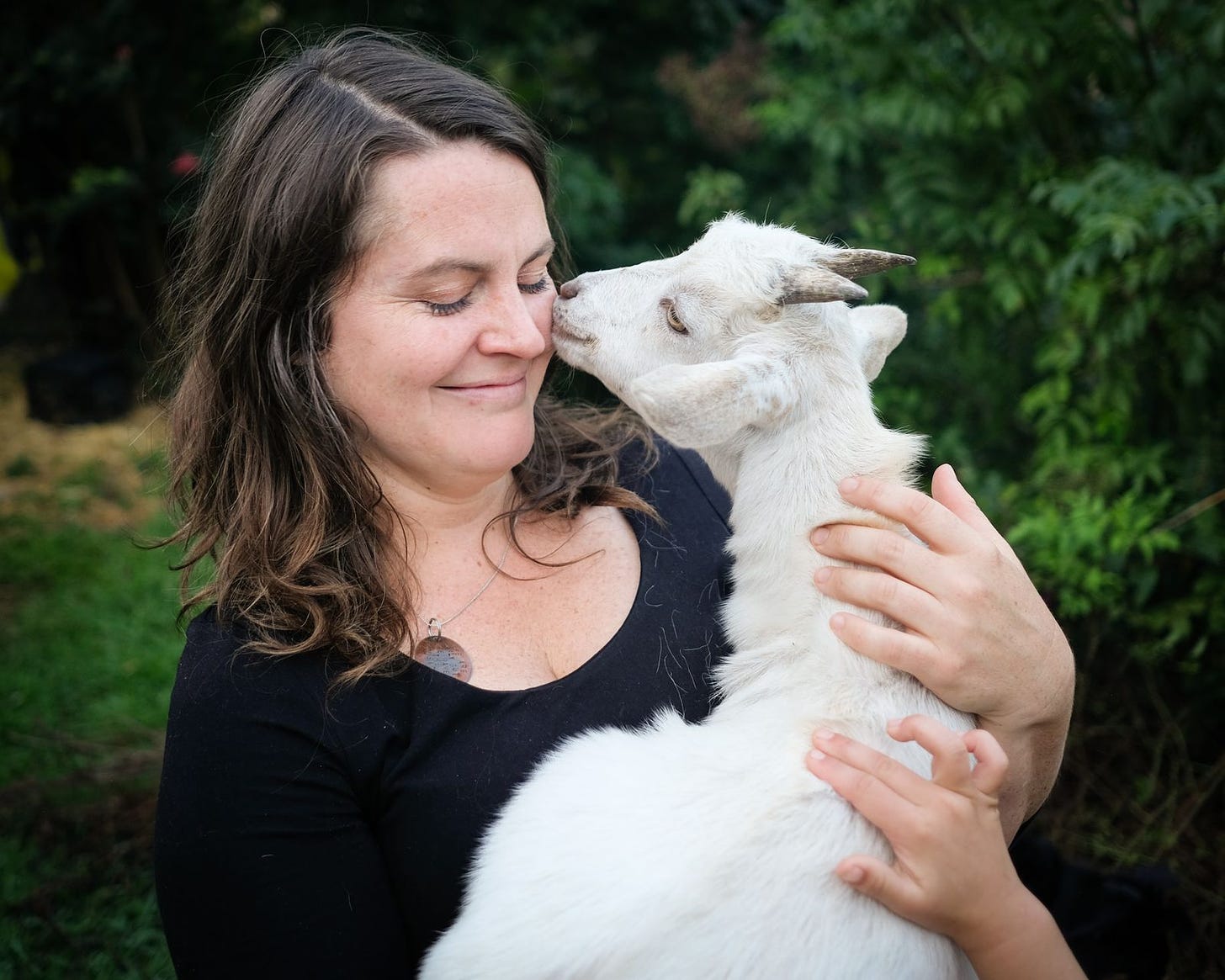 A woman stands here amid a forest scene. She is white and has brown hair, and is wearing a black shirt. It's Rachelle Barr! And her eyes are closed, and she's holding a white goat kid which is coming up to kiss her cheek. Her eyes are closed and she is smiling. Wouldn't you?