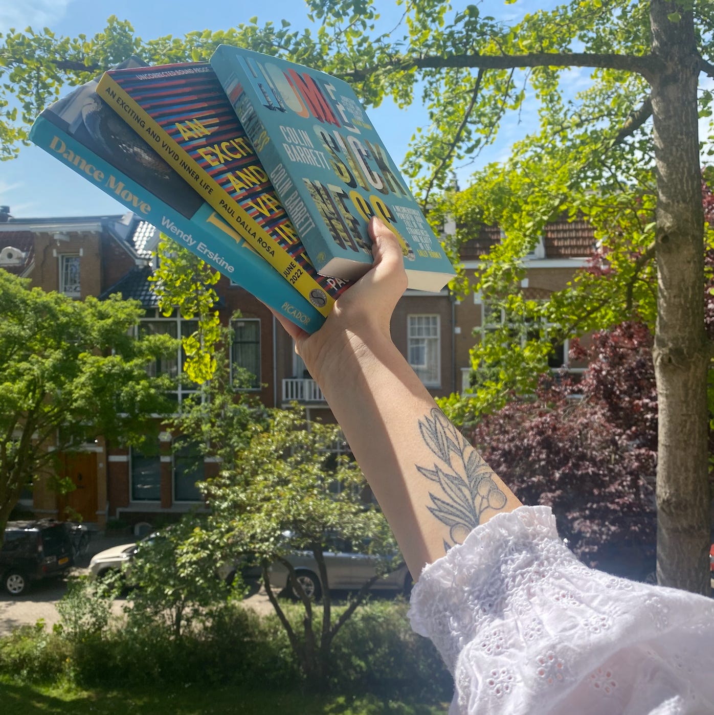hannah's tattooed arm holding three books up to the sky on her balcony 