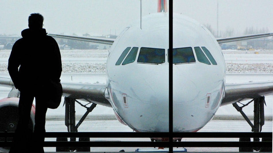 A man in an airport is looking out the window at a passenger plane. There is snow in the background.