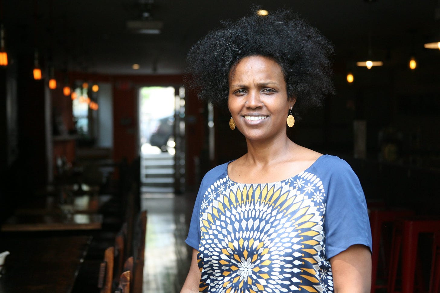 Chef Beejhy smiles wearing a blue and yellow shirt in her restaurant, Tsion Cafe