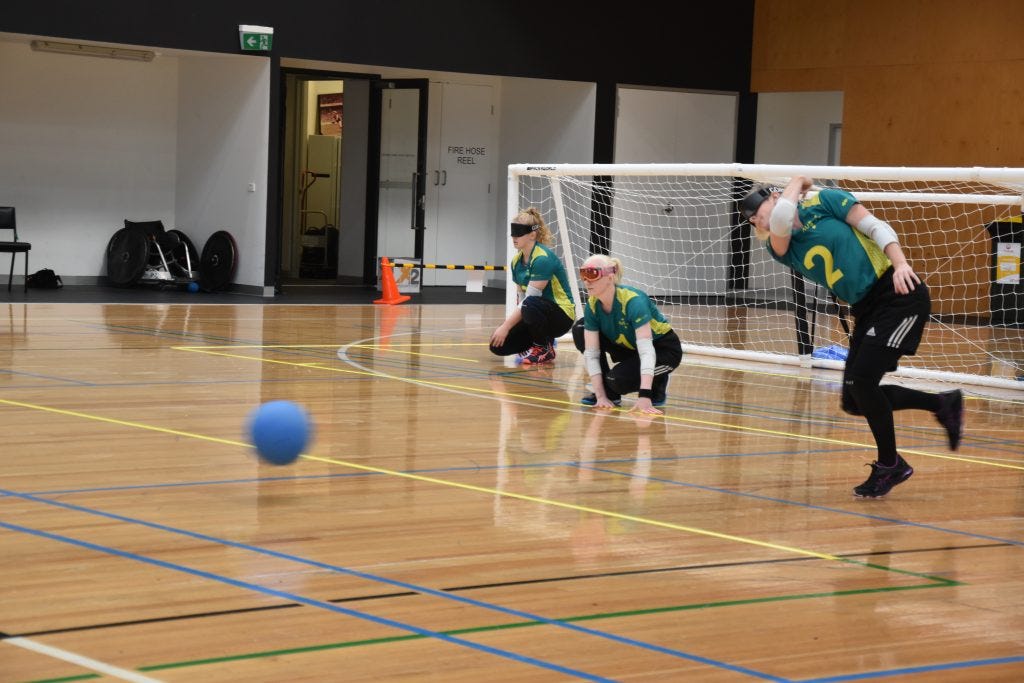 Image is of three of the Aussie Belles set up in offence positions. Two players, the middle and far left, sit crouched while the player on the far right throws the ball towards the opposition. 