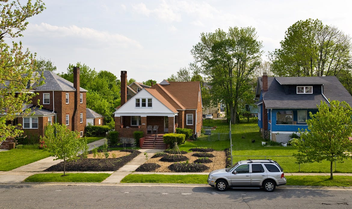 A shot of three suburban houses in a row, two with traditional ornamental front lawns, and one converted to a garden, upending the ordinary aesthetic of lot usage and suburban gardening.