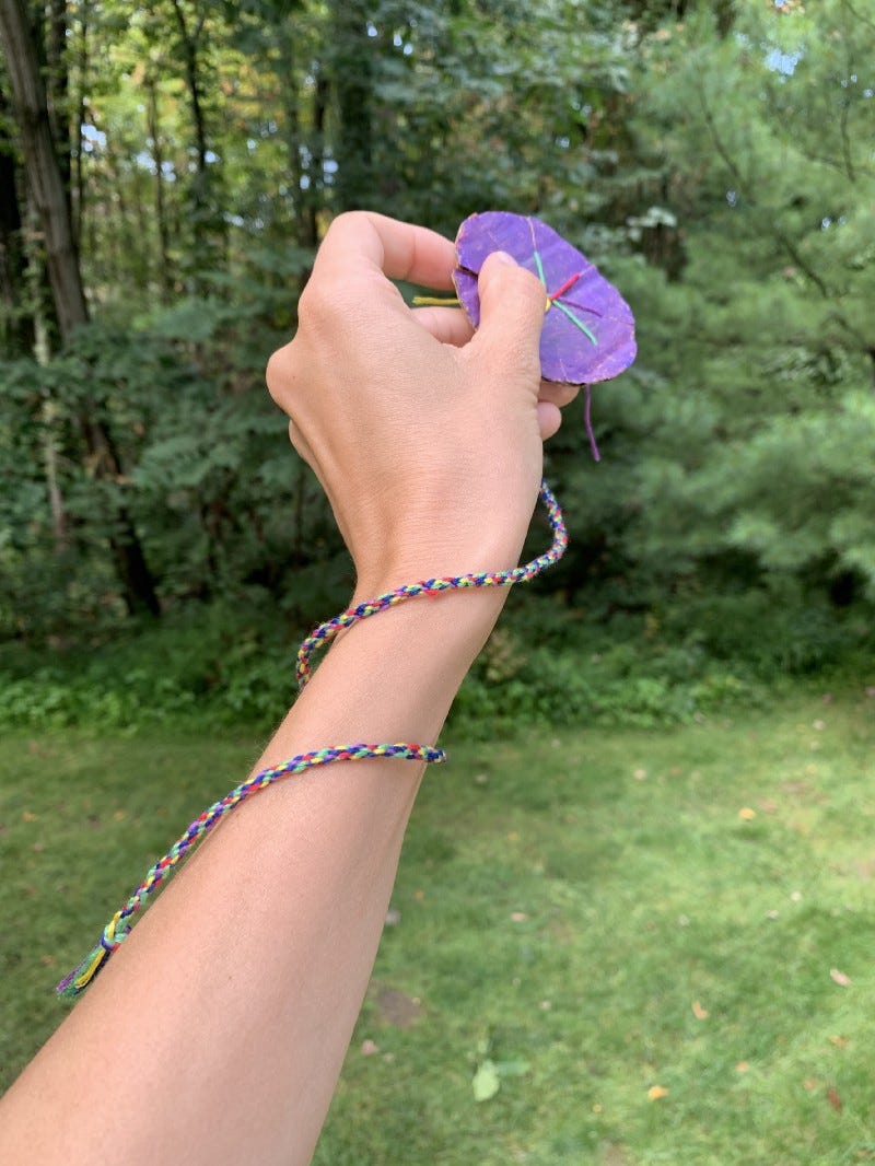 a long, multicolored friendship bracelet is held by a hand, in front of a background of green grass and trees