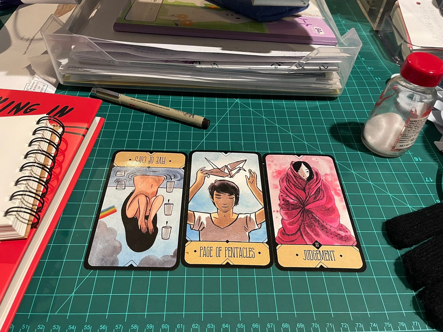 A picture of my messy desk, with a three card tarot spread in the center. The cards are five of cups reversed, page of pentacles, and judgement. Scattered around the cards is a pen, the book Crying in H-Mart, a salt shaker, gloves, and a lot of paper.