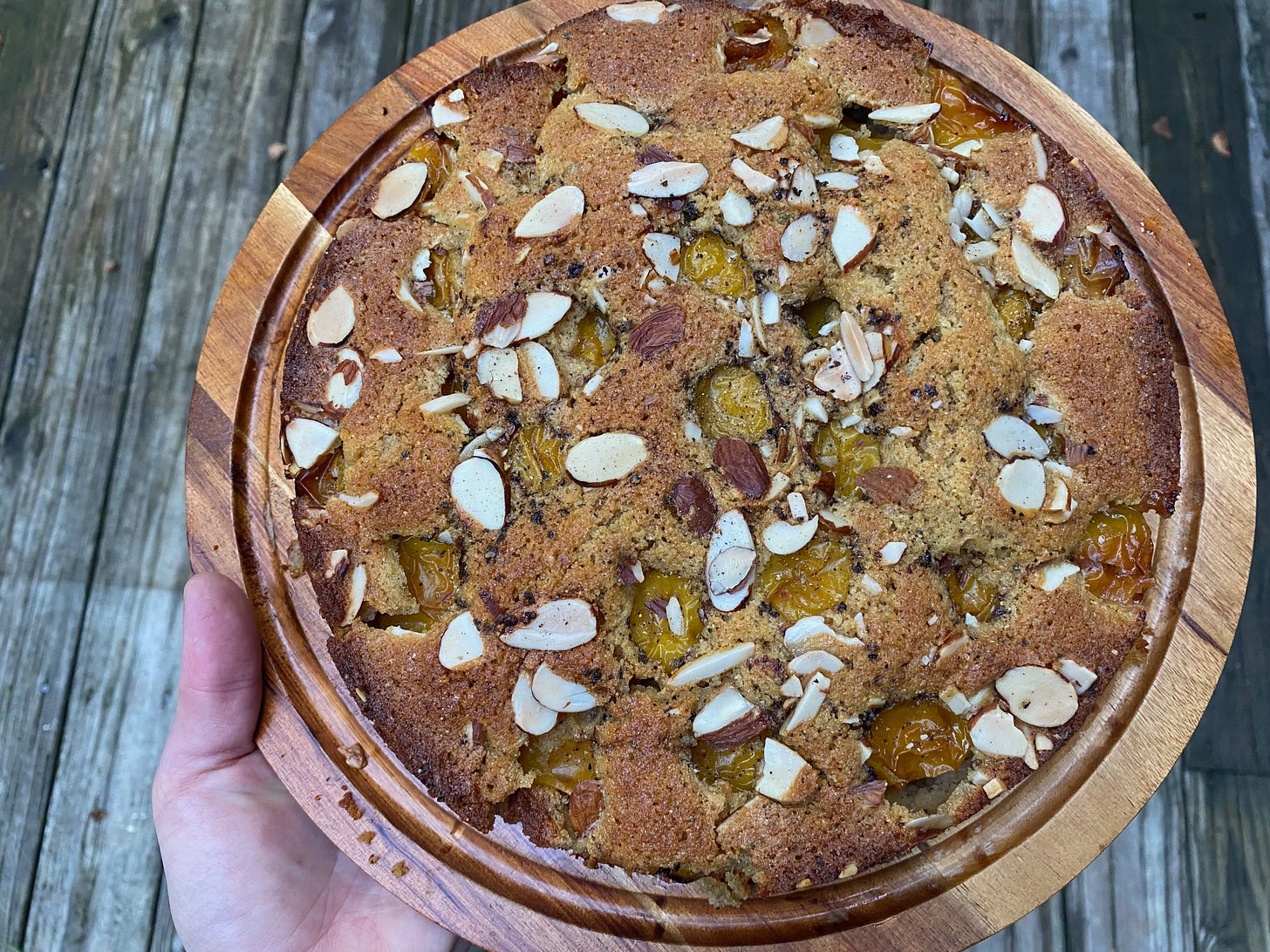 A round golden brown cake on a wooden platter. The top is studded with sliced almonds and small round golden plums.