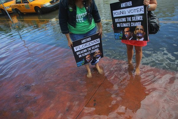 Two people stand in a flooded street. You cannot see their faces. One has a sign saying "Another Floridian breaking the silence on climate change." The other has a sign saying "Another Young Voter breaking the silence on climate change."