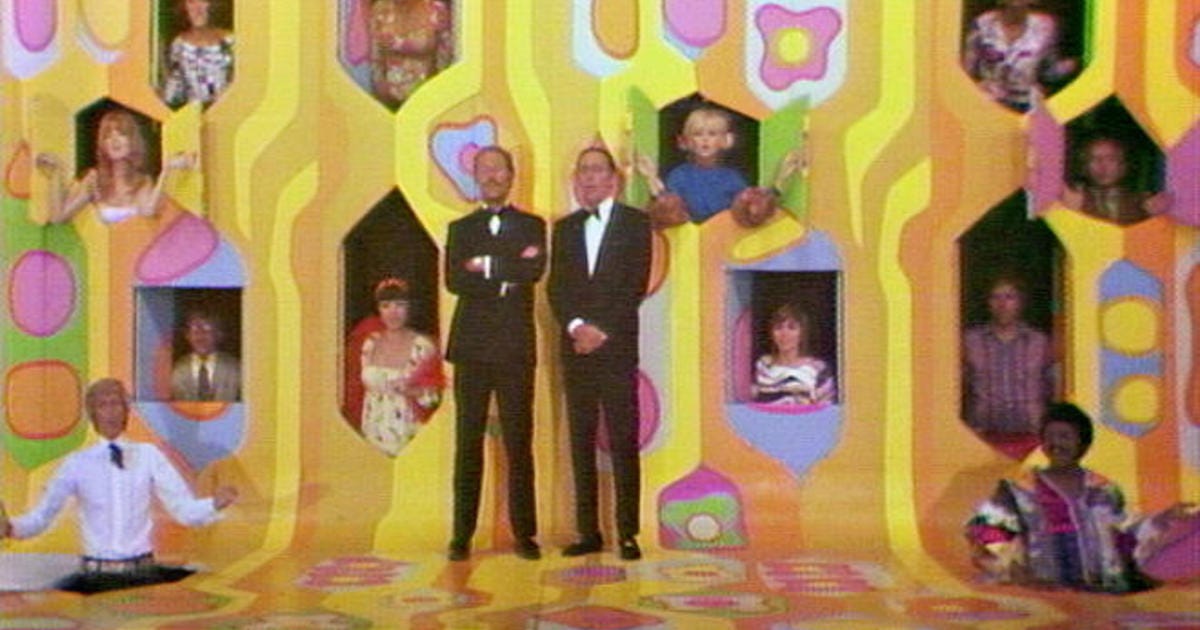 Promotional photo for "Laugh In." Two men in suits stand in a psychedelic-colored set with various windows that the cast of "Laugh In" are peeking their head through
