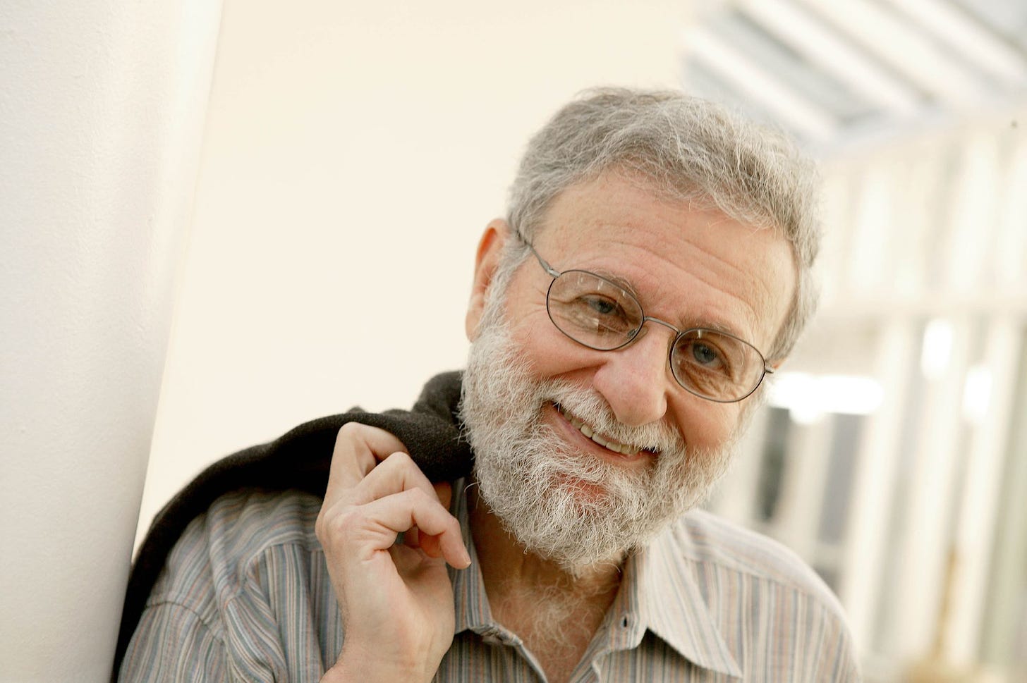 Foto mostra Don Norman, criador das expressões “Design centrado no usuário” e “Experiência do Usuário” em pose informal, de óculos, barba, sorrindo, recostado em uma parede e segurando um casaco no ombro.