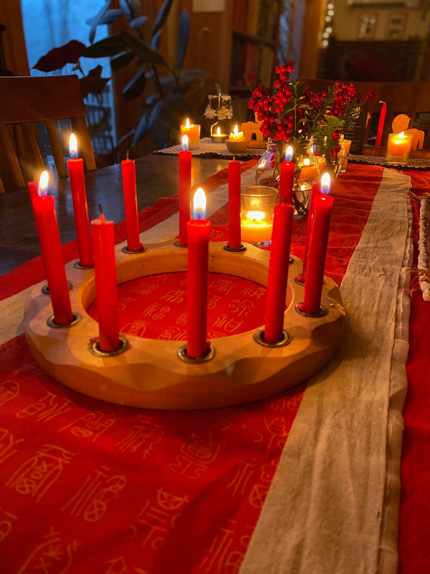 A circular wooden candle holder full of lit red candles sits on a red runner on a wooden table. More lit candles and jars of winterberry are arranged behind it.