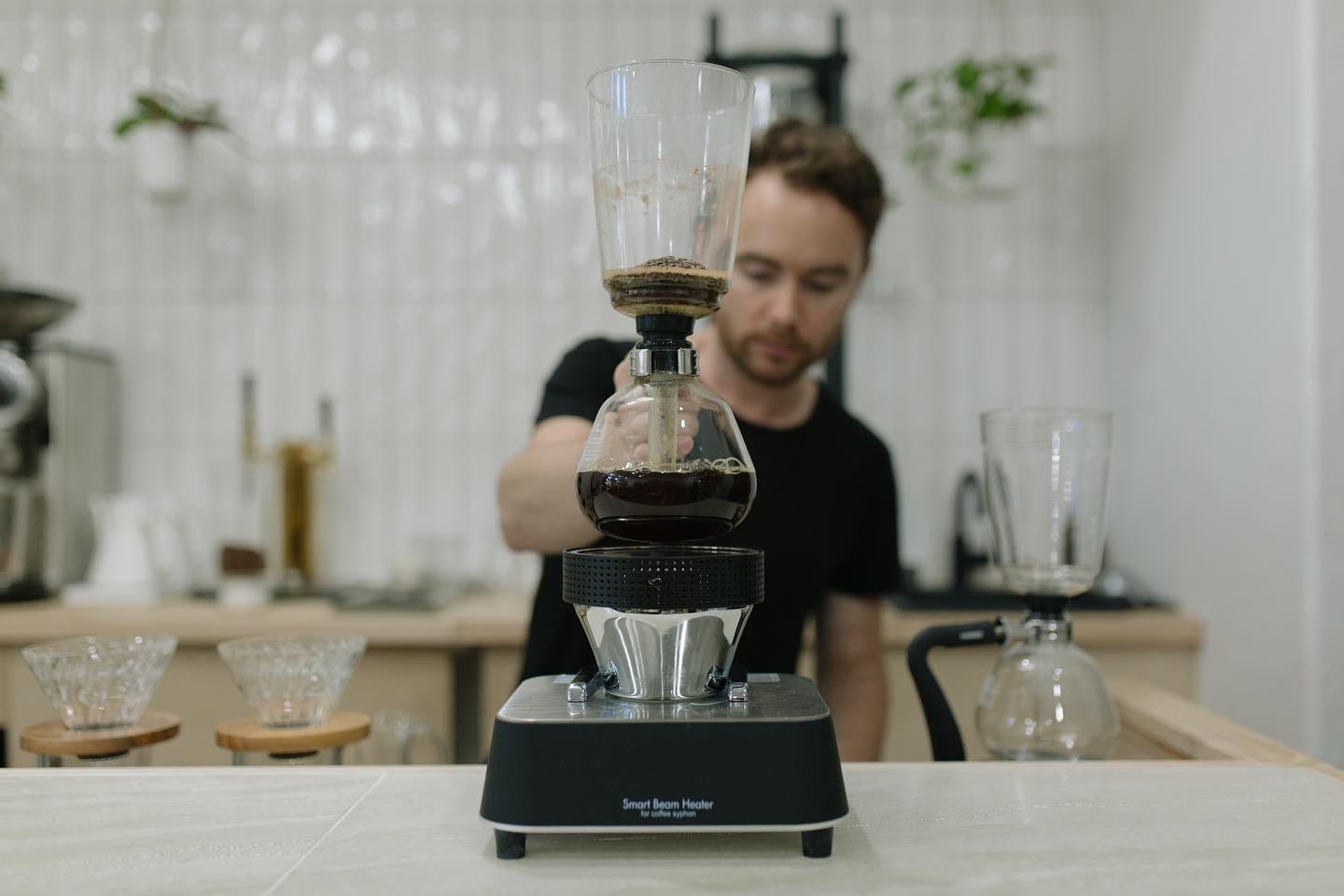 A close-up photo of a siphon coffee brewer in use. A globe half-filled with coffee rests on a black heating device. A tube runs from the globe to an upper glass chamber with the spent coffee grounds. A barista is blurred in the background against a white subway tile wall.