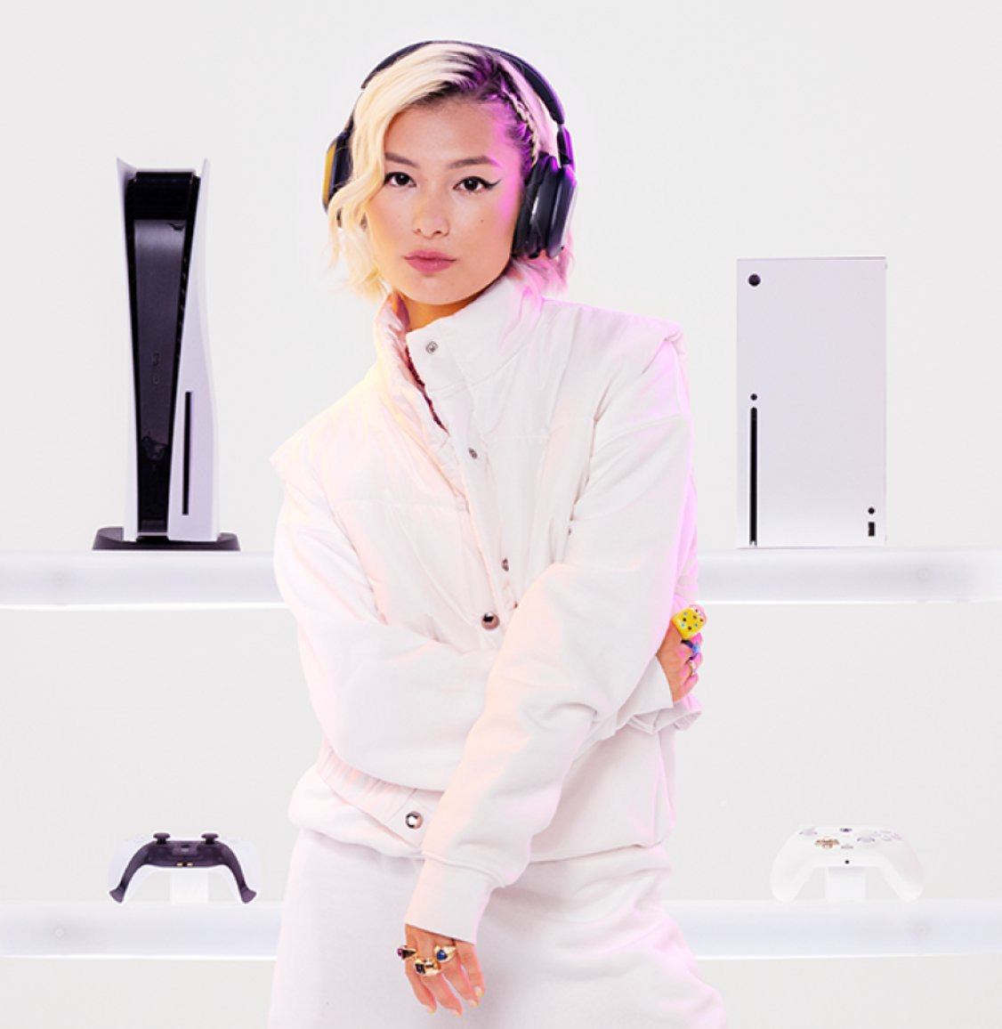 A white Xbox Series X and PS5 in the background of an Asian woman wearing headphones dressed in white