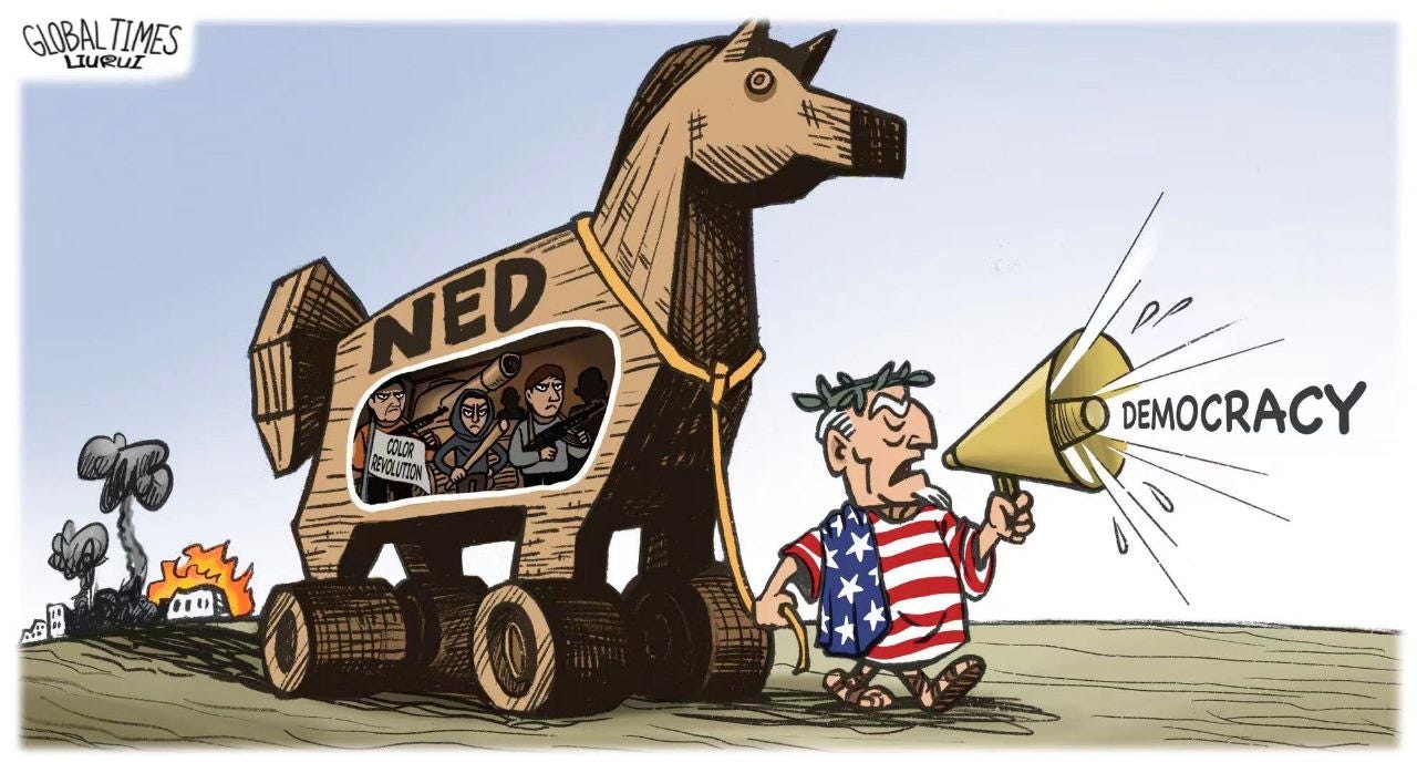 A man in an Amerikan flag toga is holding a megaphone and yelling "Democracy". He is pulling a large wooden horse that says NED (National Endowment for Democracy) on the side. Part of the belly of the horse is cut-away to reveal militants inside with weapons and a sign that says "color revolution". Behind the horse is the remnants of a city that is burning.