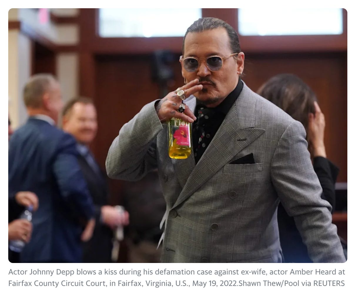 Color photo of Johnny Depp in a courtroom with tied back dark hair, wearing a tweed suit with black shirt and tie, holding his right hand up with two fingers before his mouth. He is wearing three rings on that hand. Caption says: “Actor Johnny Depp blows a kiss during his defamation case against ex-wife, actor Amber Heard at Fairfax County Circuit Court, in Fairfax, Virginia, U.S., May 19, 2022”