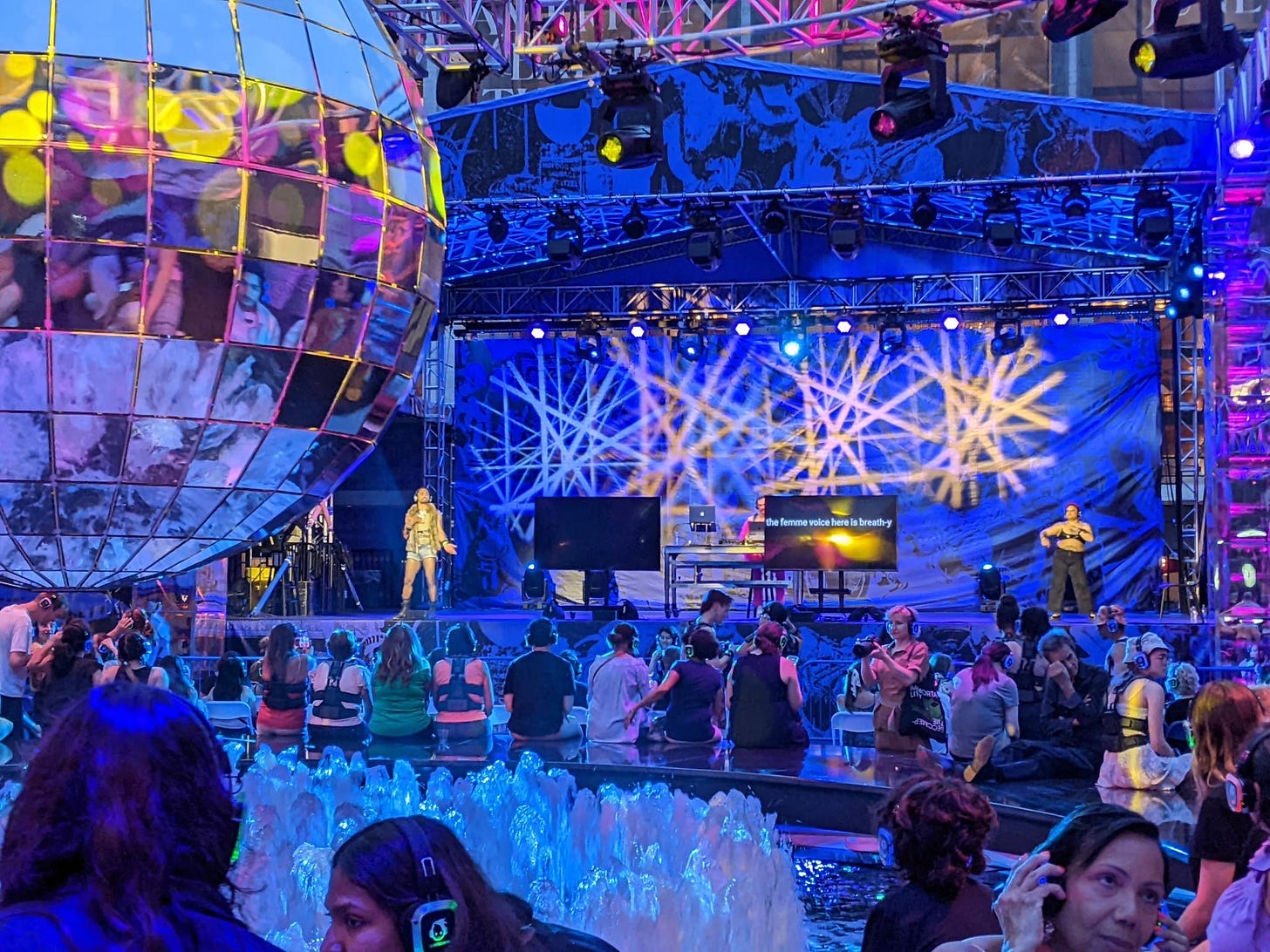 The outdoor dance floor at Lincoln Center is bathed in blues and purples. A giant 10 foot disco ball hangs over babbling fountains. People wear headsets, lounging. There is a big stage with an ASL interpreter, dancer, and DJ in between two screens. One says ‘the femme voice here is breath-y.”
