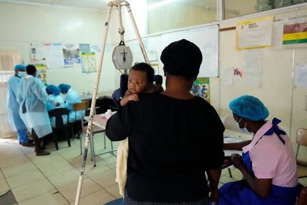 A view from behind of a mother, in a black shirt and black cap, holding her baby at her shoulder, while a medical worker takes notes wearing a blue apron, blue cap and blue face mask and pink shirt. In front of the mother is a large scale for weighing the baby, and in the background, a group of health workers in blue P.P.E. are talking.
