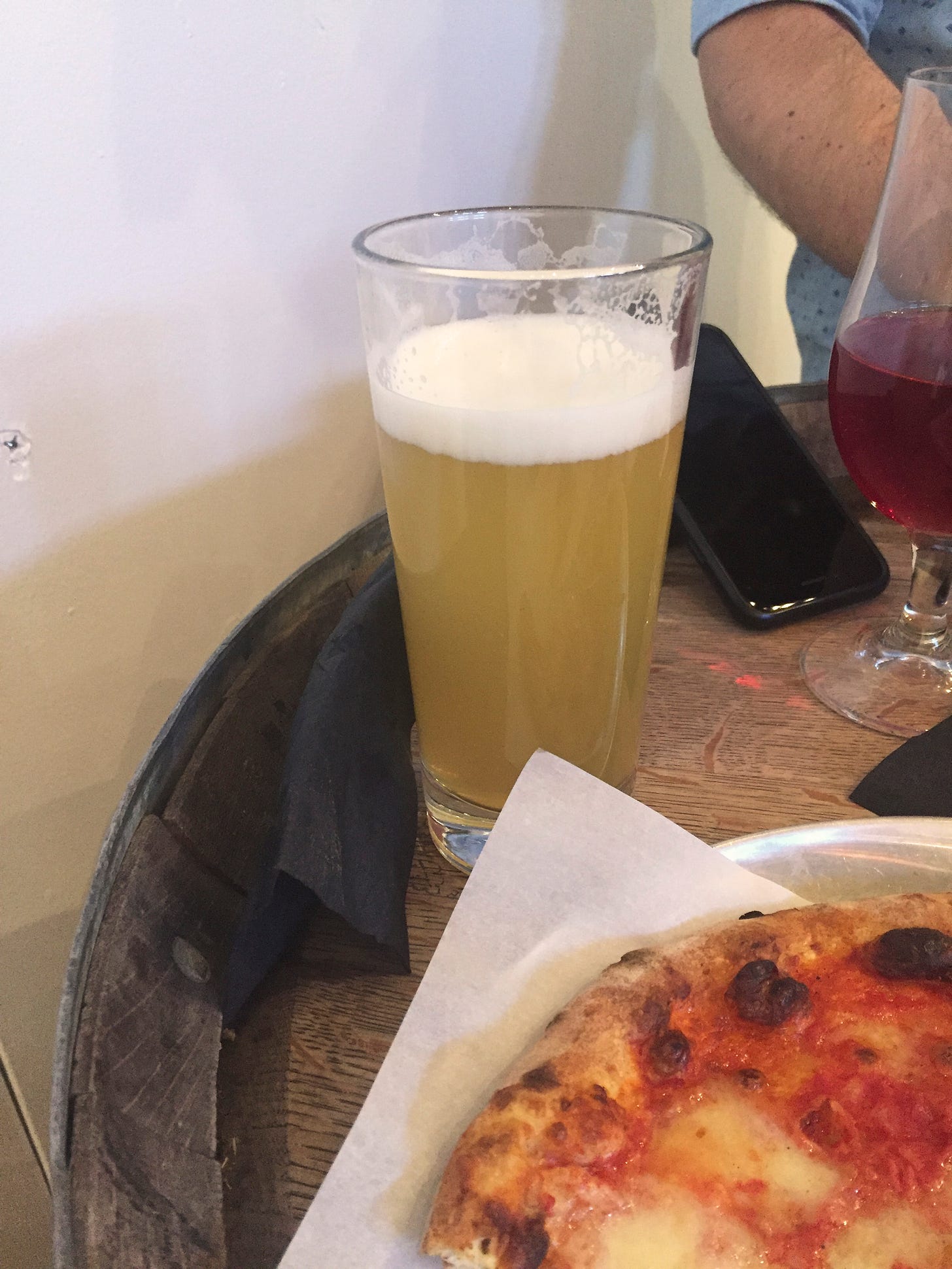 A glass of hazy IPA sits on top of a wooden barrel. At the corner of the frame in front of it, part of an Italian pizza is visible. In the background is a tulip glass of pinkish raspberry sour ale.