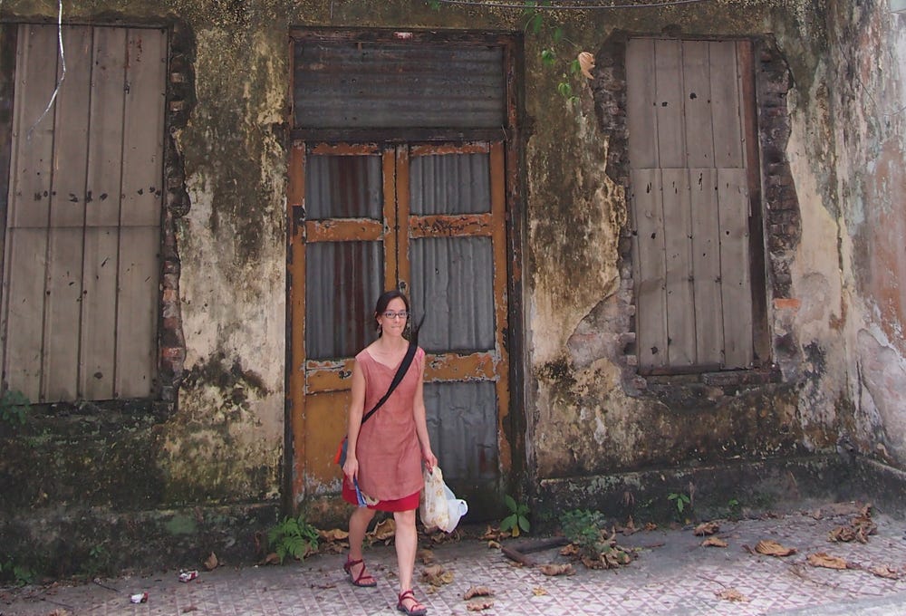 A light skinned, mixed race woman in a red top and skirt walks away from a decaying building. The doors and windows are boarded up and the plaster is crumbling and moldy.