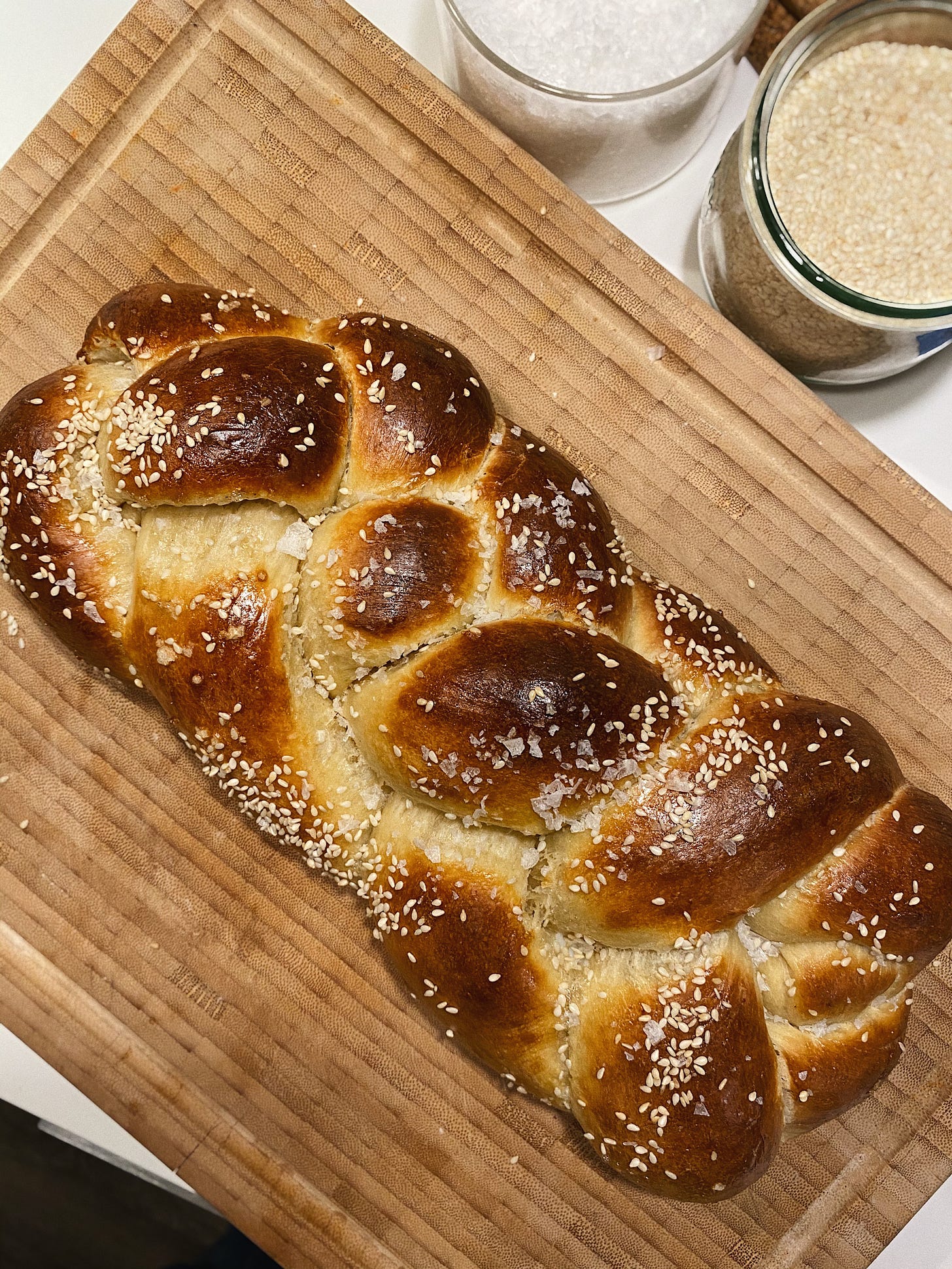 a braided loaf of challah bread sprinkled with sesame seeds and sea salt, on top of a light bamboo cutting board with two jars, one filled with sesame seeds and the other with salt.