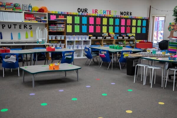 In a classroom, chairs, stools and tables with school supplies on them sit before a wall lined with colored papers and the label “Our Brightest Work.”