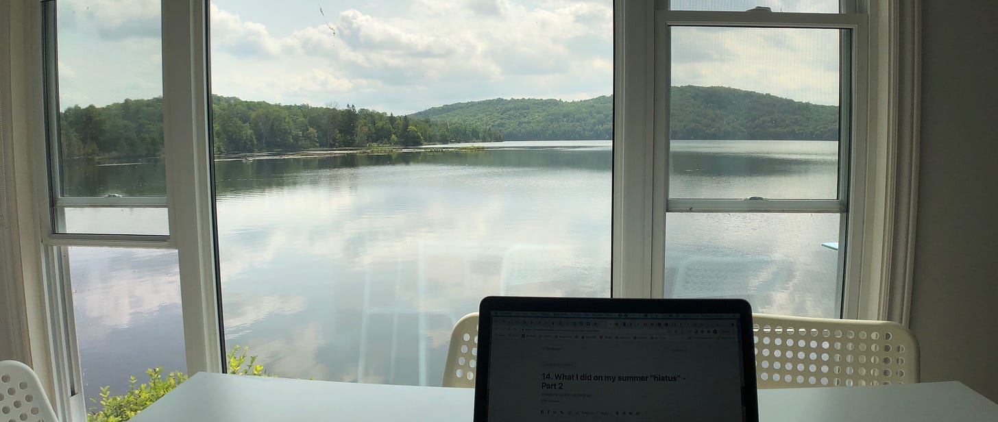 A computer on a table in front of a large window. Beyond the window is a wide view of a placid lake and green, rolling hills in the background. 