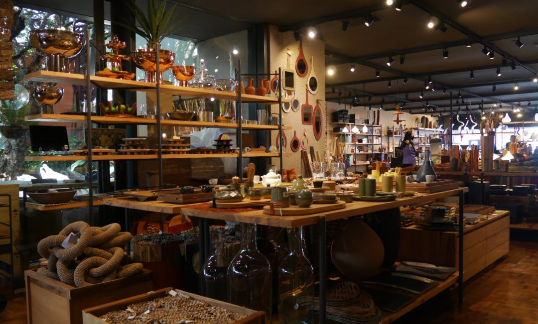 Interior de loja de produtos de design brasileiro, com prateleiras ao redor de uma grande mesa de madeira com produtos expostos.