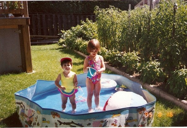 Childhood photo of Cassandra and her sister in a kiddy pool in their grandparents' backyard, next to the garden full of blooming tomato and basil plants