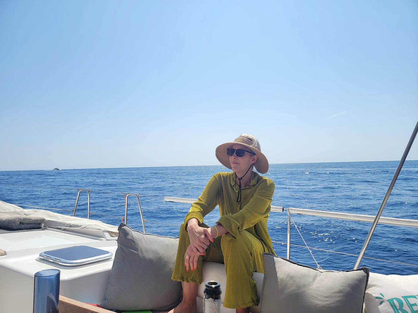 The author sits on a sailboat in a lime pants and shirt, sunglasses, and a sun hat. A cobalt blue ocean stretches behind her.
