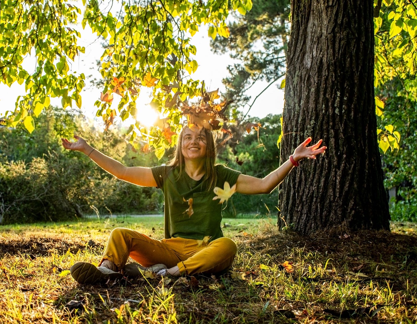 A woman smiles and throws a piles of leaves above her head in the bright sunlight of a park.