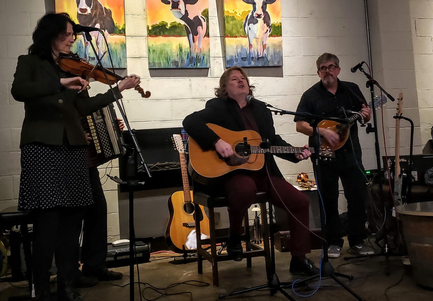Violin, accordion and mandolin players stand around a seated guitarist/ singer. Cool paintings of cows on the wall behind them. 