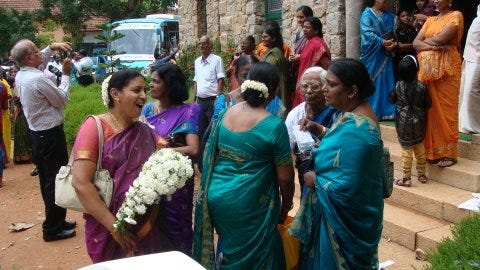 Image: Women in saris, with strings of jasmine in their hair, confer seriously near the steps of a church. Behind them, a grey-haired man leans back to carefully photograph smiling guests. A bright blue tourist bus, perhaps intended for the bride’s aunts and cousins, is parked incongruously near the hedge and old stone masonry.