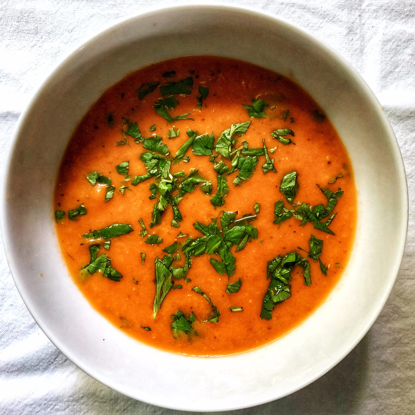 Tomato soup with cilantro sprinkled on the surface. The soup is in a white bowl, which is placed on a white fabric background.