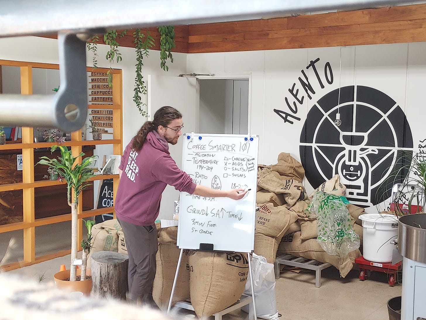 A pony-tailed man writes coffee chemistry facts on a white board set up in front of a bunch of green coffee in burlap sacks.